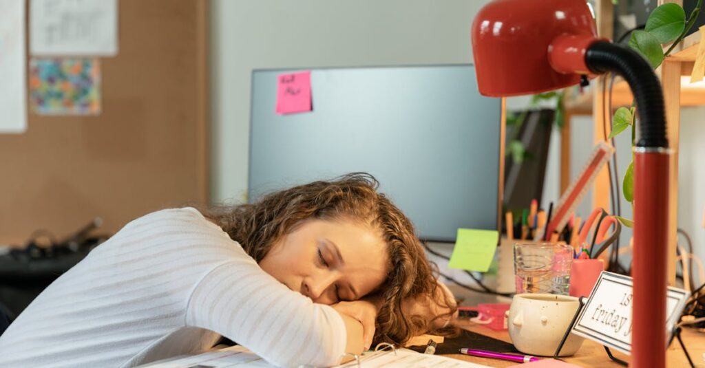 A woman sleeping at her office desk, surrounded by work documents and a red desk lamp.